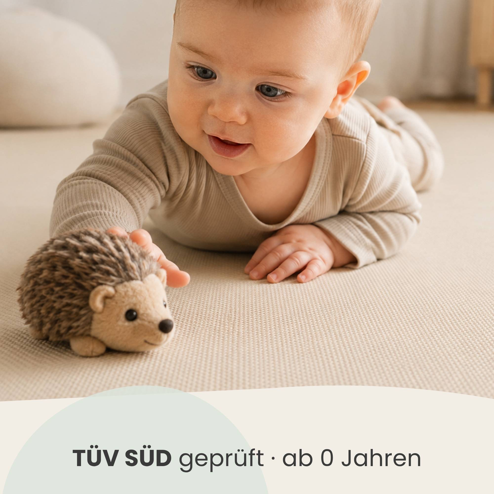 Baby liegt auf einer Matte und greift nach einem Spielzeug-Igel, mit Kennzeichnung „TÜV SÜD geprüft · ab 0 Jahren