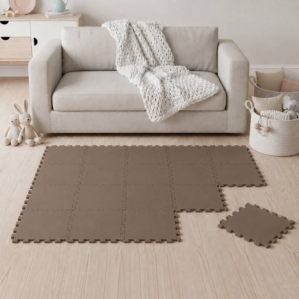 Ein minimalistisches Wohnzimmer mit einem beigefarbenen Sofa und einer Decke, das braune Schaumstoff-Bodenfliesen teilweise zusammengesetzt zeigt und eine lose Fliese auf dem Boden hat.