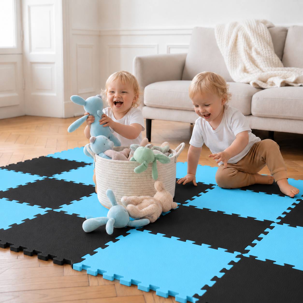 Zwei Kleinkinder spielen auf einer schwarzblauen Schaumstoffmatte, lachen mit Plüschhäschen neben einem Spielzeugkorb in einem hellen Wohnzimmer.