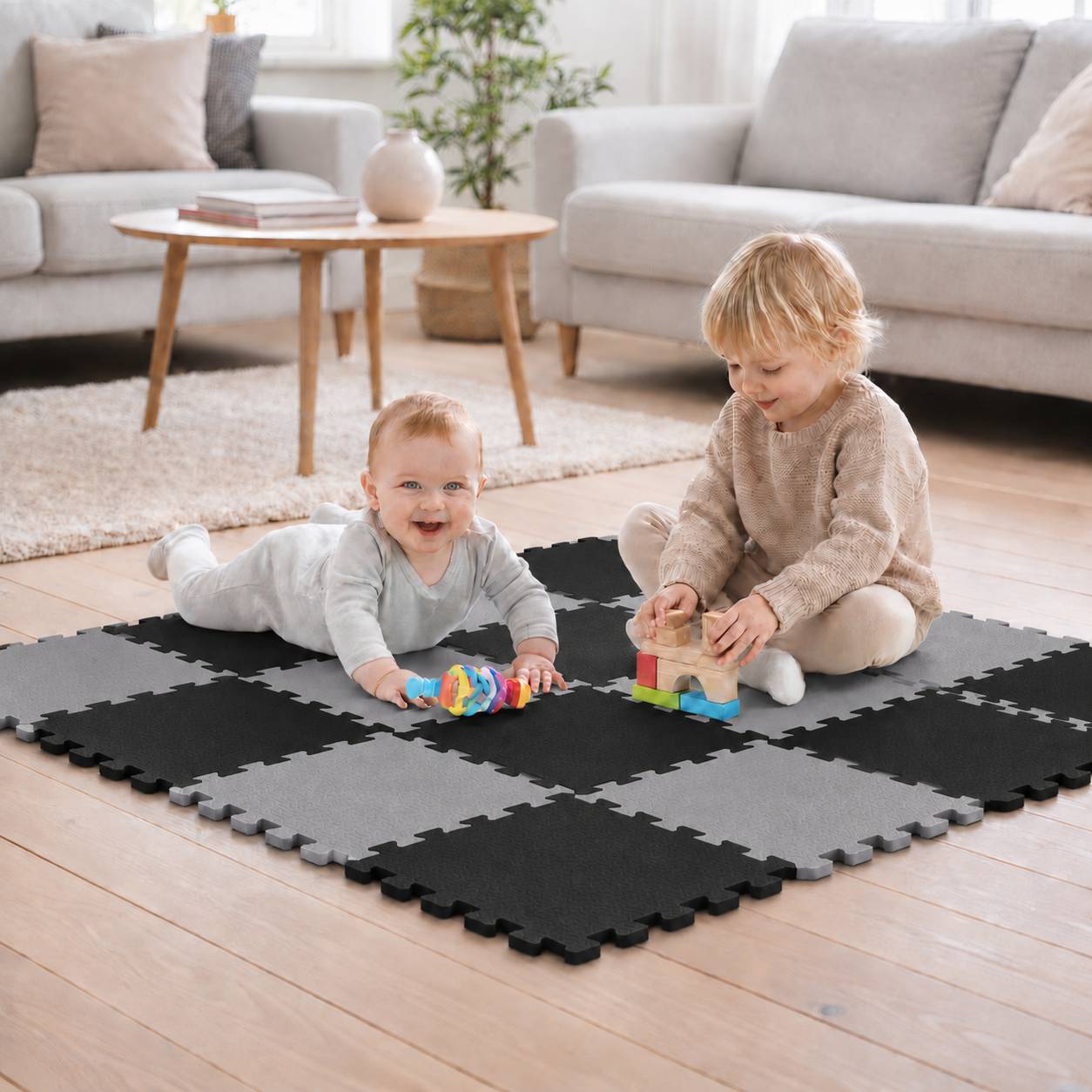 Zwei kleine Kinder spielen mit Spielzeugen auf einer schwarzen Schaumstoffmatte in einem hellen Wohnzimmer.