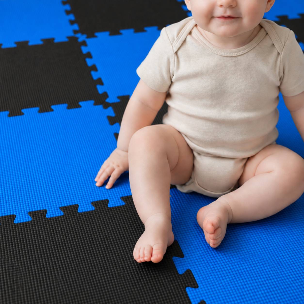 Ein Baby in einem beigefarbenen Strampler sitzt auf blau-schwarzen ineinander greifenden Schaumstoff-Spielmatten.