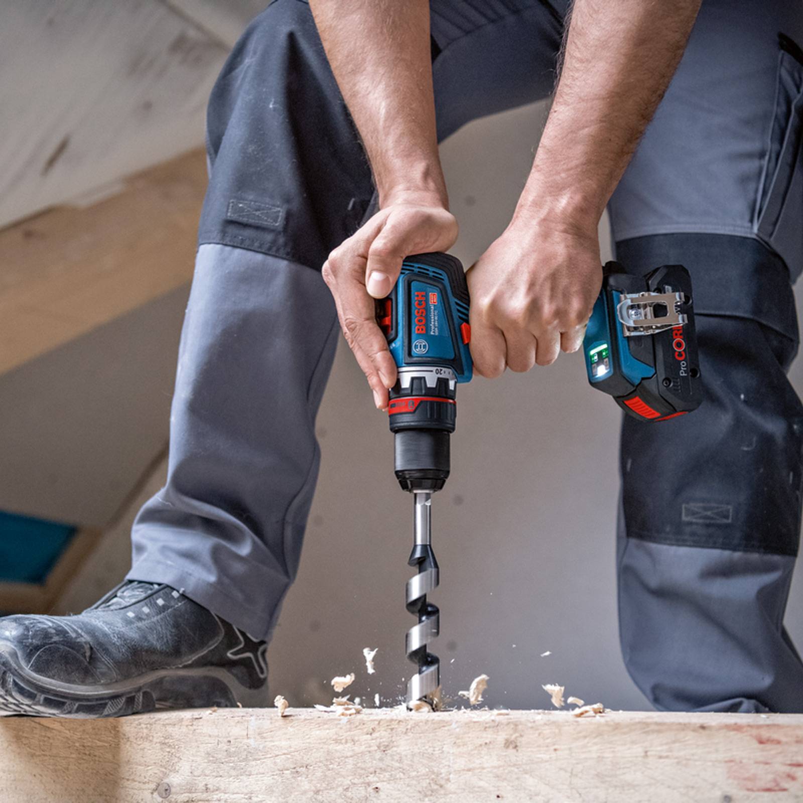 Eine Person benutzt einen Bosch-Akkuschrauber, um ein Loch in einen Holzbalken zu bohren, trägt Sicherheitsausrüstung und befindet sich auf einer Baustelle.