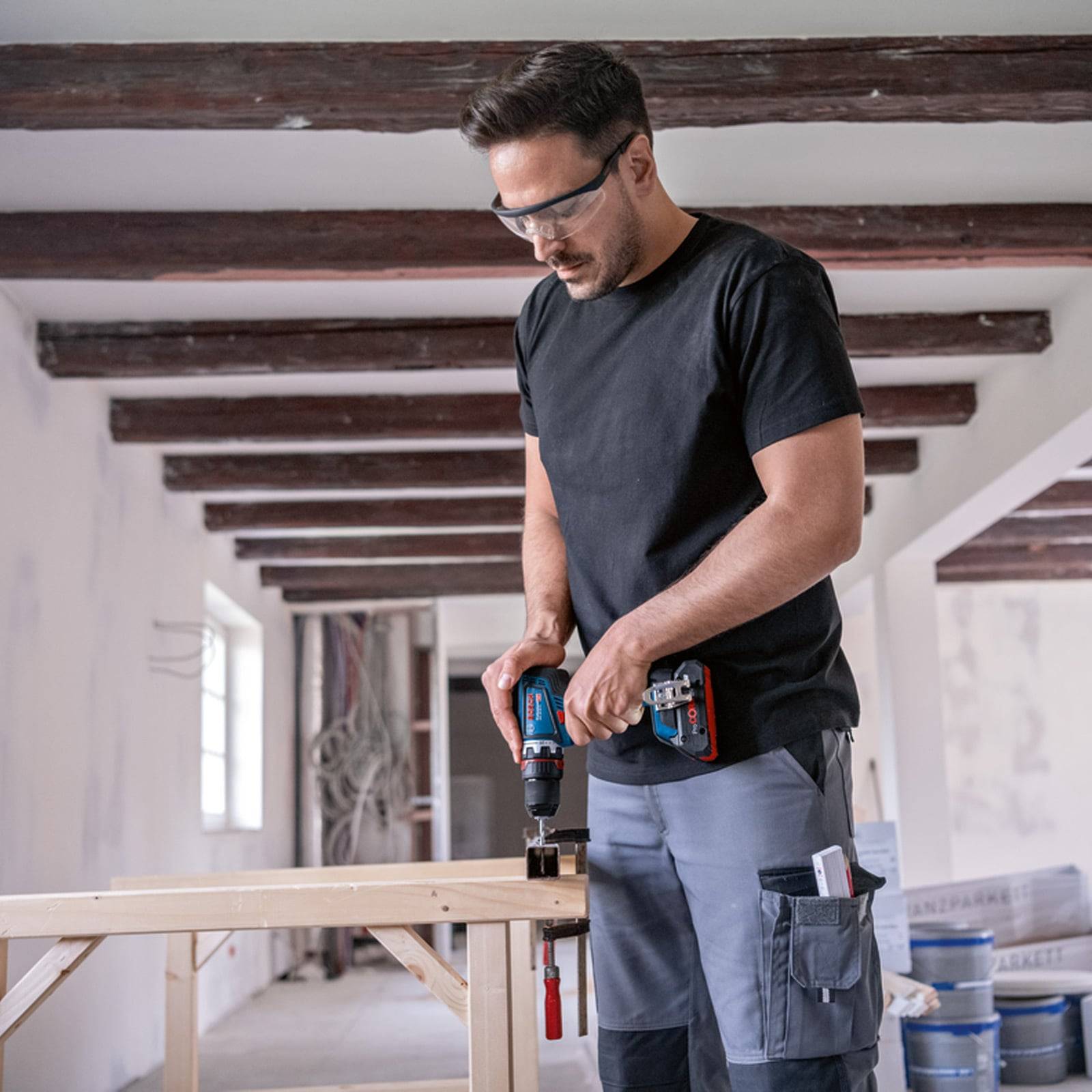 Ein Mann, der in einem teilweise fertiggestellten Raum eine Bohrmaschine auf einem Holzbrett verwendet und dabei Sicherheitsbrille und lockere Arbeitskleidung trägt.