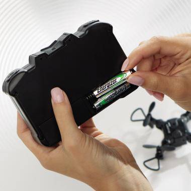 Hände führen zwei Energizer AA-Batterien in ein schwarzes Batteriepack oder Gerät ein, mit einem kleinen ferngesteuerten Hubschrauber im Hintergrund.