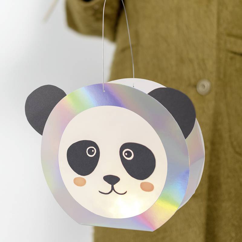 Eine Person hält eine Laterne in Form eines Pandagesichts aus irisierendem Papier, in der ein Licht von innen leuchtet, vor einem unscharfen Hintergrund.