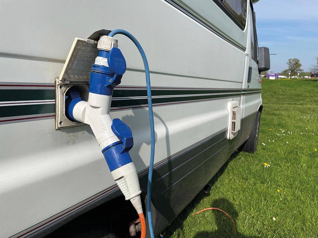 Blau-weißer Adapter, der in eine Stromanschlussbuchse eines Wohnwagens gesteckt ist, mit Kabel, das neben dem geparkten Fahrzeug auf dem Gras zum Boden verläuft.