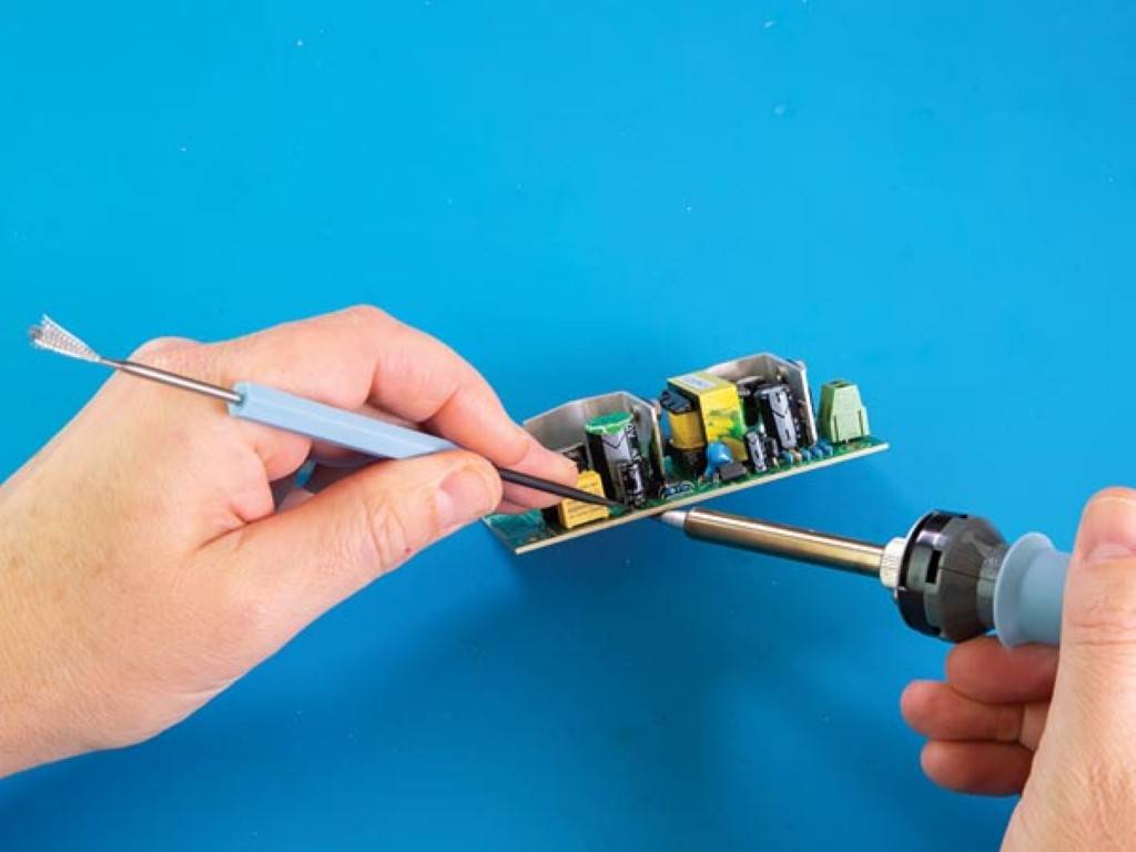 Hände halten eine Leiterplatte, während ein Lötkolben verwendet wird, um elektronische Komponenten auf blauem Hintergrund zu reparieren.