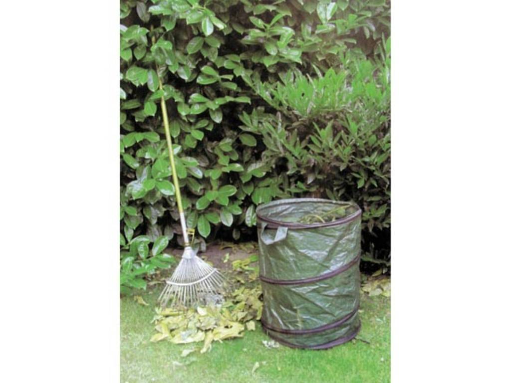Gartenrechen, der neben einem grünen Maschengartenabfallsack lehnt, der mit Blättern gefüllt ist, auf einem Rasen, vor dichten Sträuchern.