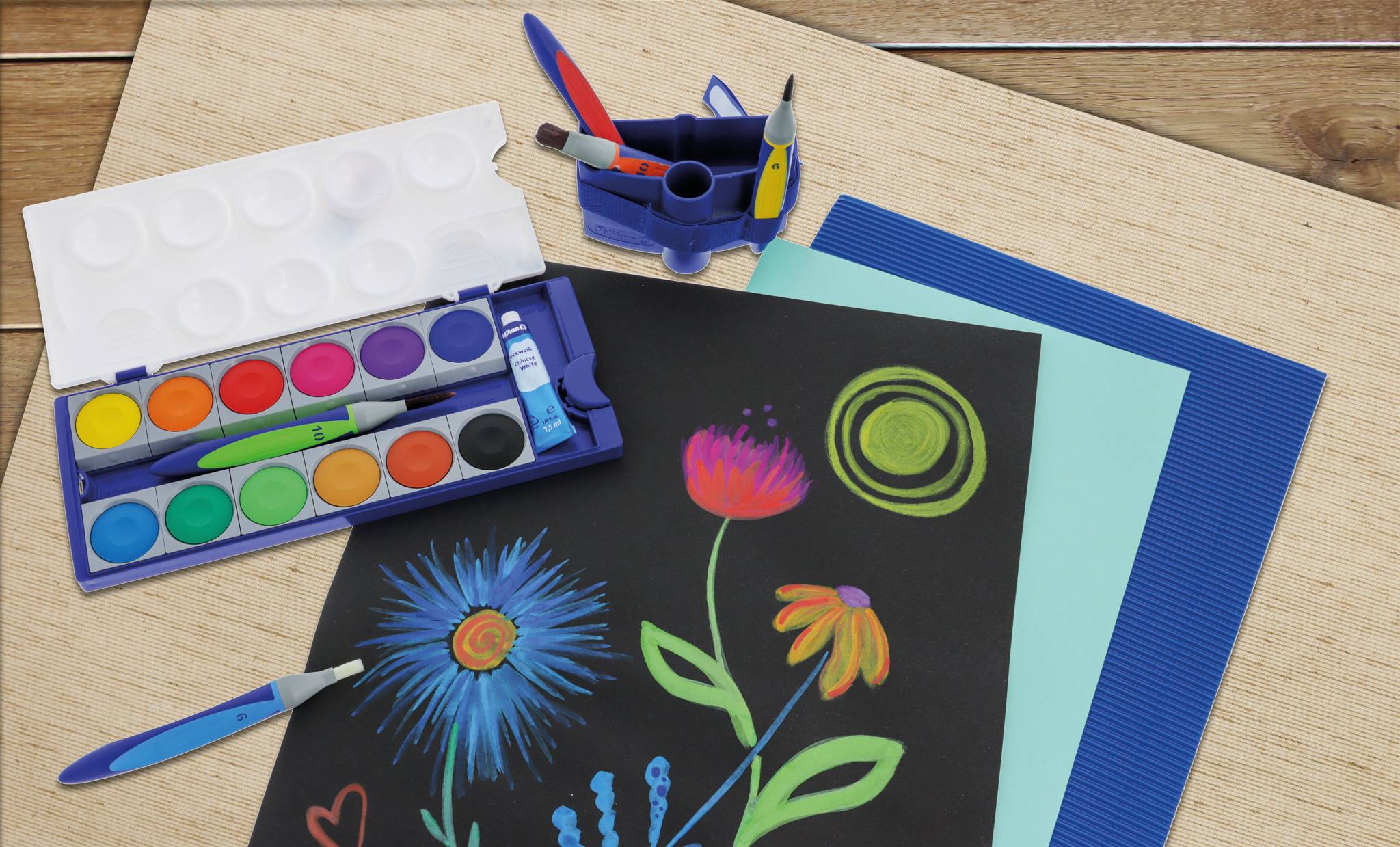 Kunstmaterialien auf einem Schreibtisch: ein offenes Aquarellmalkasten-Set, ein Pinsel, farbiges Papier und ein schwarzes Blatt mit leuchtend bemalten Blumen und Kreisen.