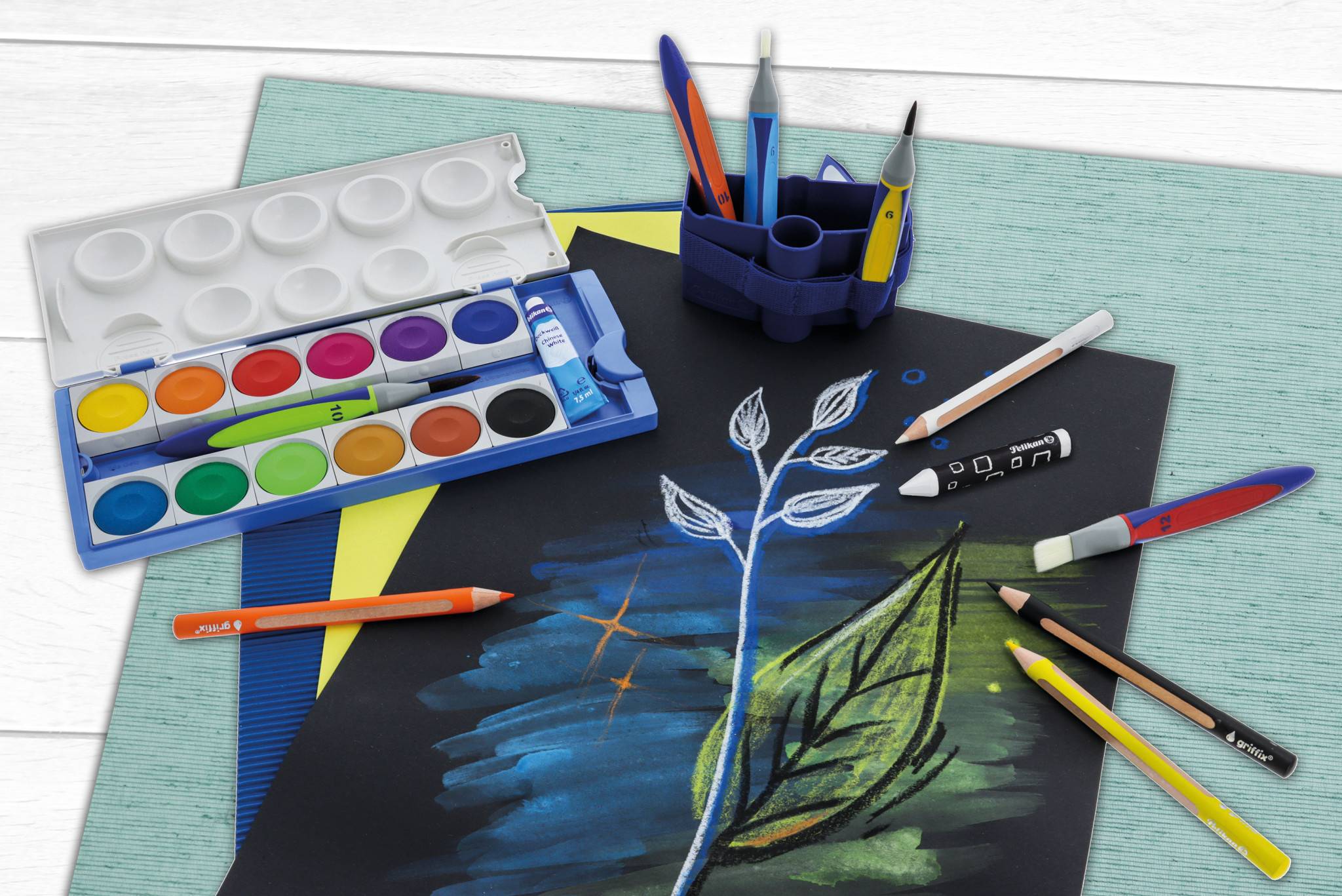 Kunstzubehör auf einem Schreibtisch mit einer Aquarellpalette, Bleistiften, Markern und einer Zeichnung eines Blattes auf schwarzem Papier.