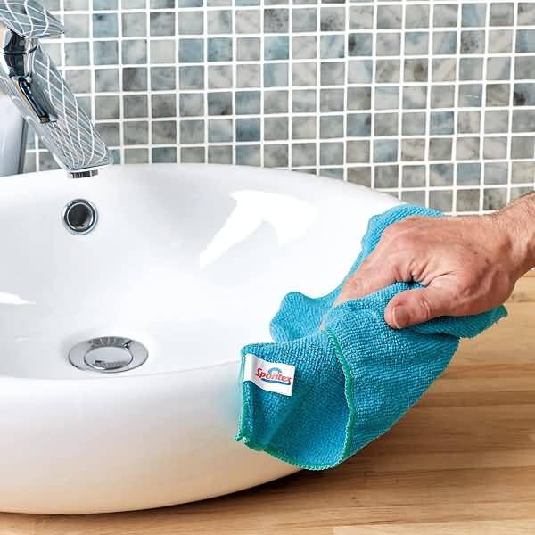 Hand wischt ein weißes Badezimmerbecken mit einem blauen Reinigungstuch neben einem Chromwasserhahn und einer gefliesten Rückwand ab.