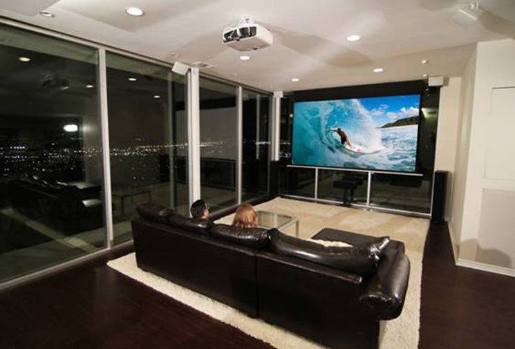 Modernes Heimkino mit zwei Personen auf einem schwarzen Sofa, die ein Surf-Video auf einem großen Bildschirm ansehen, in einem Raum mit Glaswänden und Stadtlichtern außen.