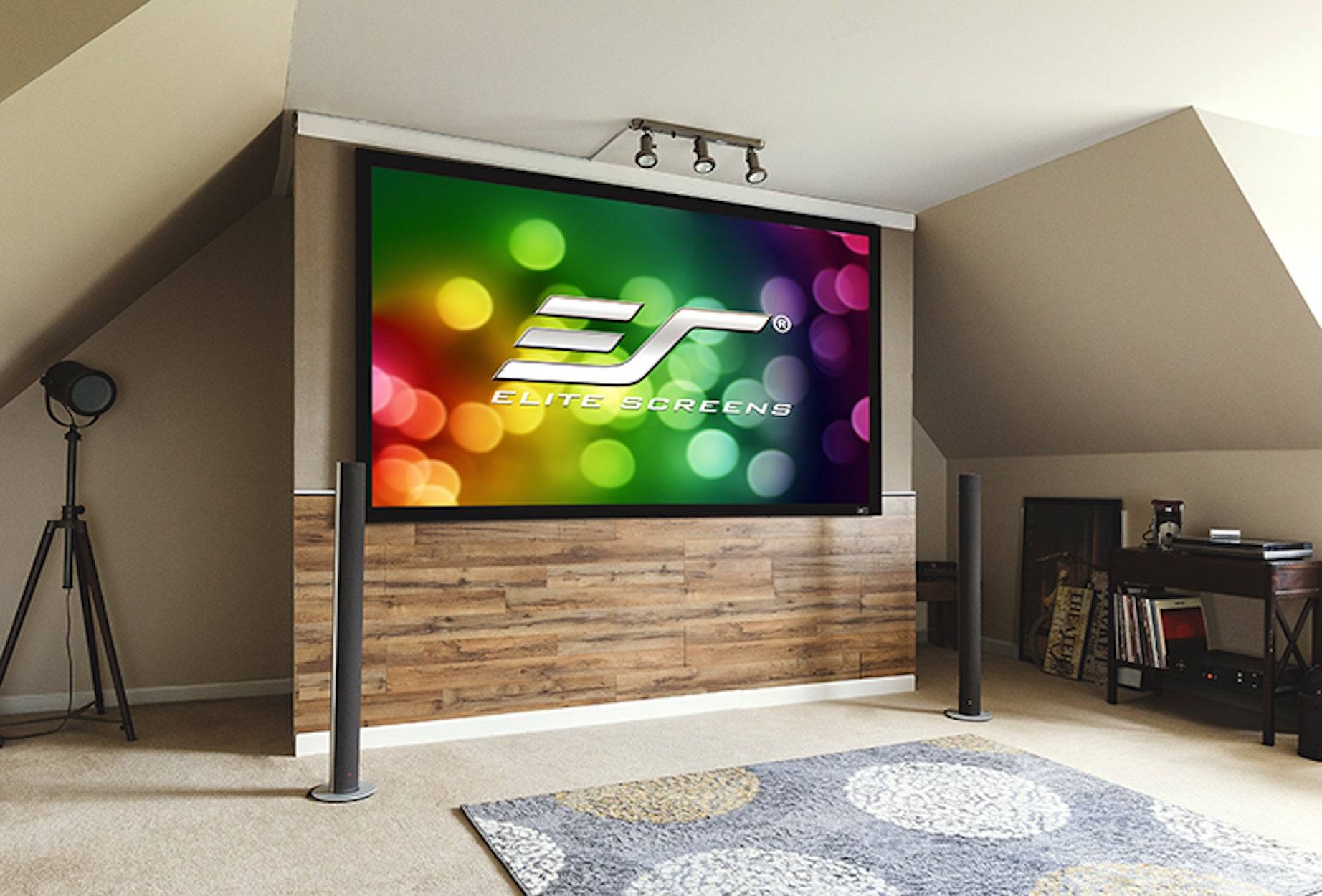 Ein großer Heimkino-Projektionsbildschirm auf einer erhöhten Plattform zeigt das Logo "Elite Screens"" und einen bunten Bokeh-Hintergrund in einem Dachboden-ähnlichen Raum.