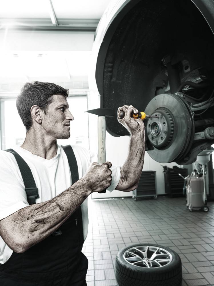 Ein Mechaniker arbeitet an der Bremsscheibe eines Autos in einer Werkstatt und benutzt dabei einen Schraubenschlüssel. Die Umgebung ist industriell geprägt, mit Werkzeugen, die im Hintergrund sichtbar sind.