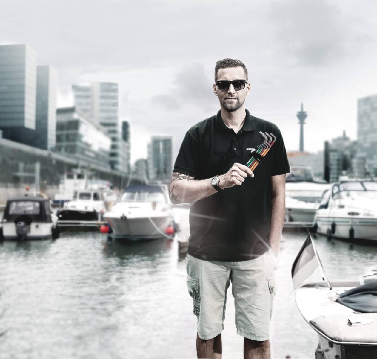 Eine Person mit Sonnenbrille steht selbstbewusst vor einer Marina; moderne Stadtlandschaft mit Gebäuden und Booten im Hintergrund.