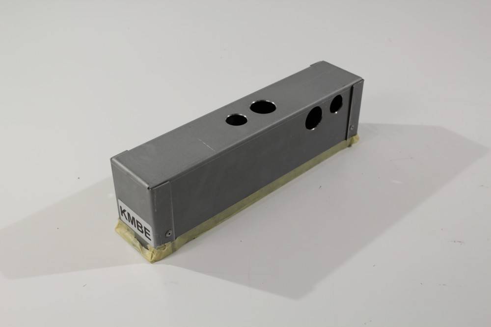 Eine graue rechteckige Metallbox mit drei kreisförmigen Löchern auf der Oberseite und einem Aufkleber mit der Aufschrift 'KMBE' an der Seite, die auf einer weißen Oberfläche steht.