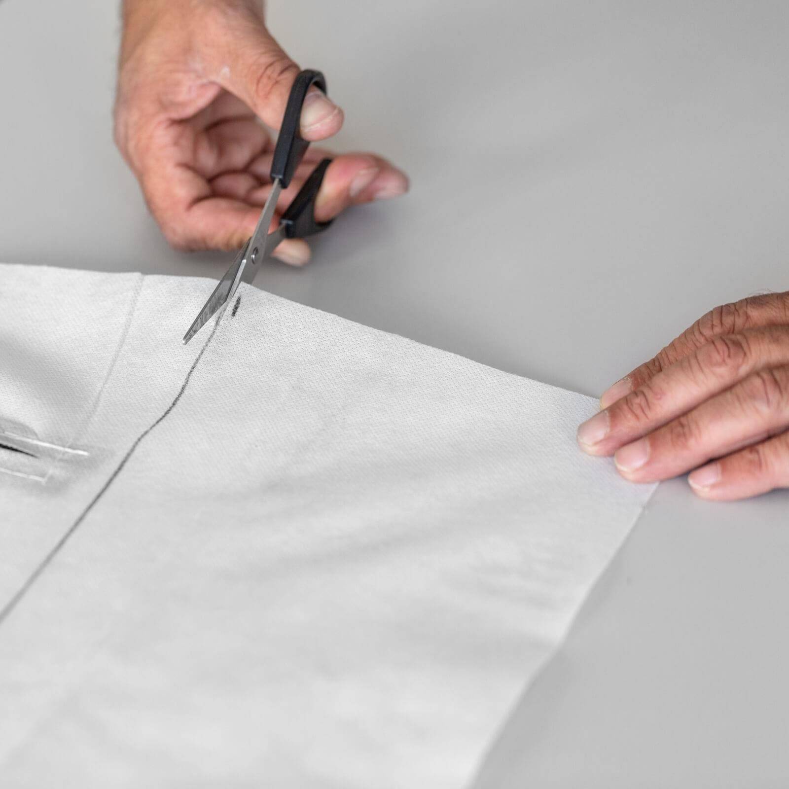Hände, die eine Schere verwenden, um ein weißes Stück Stoff oder Papier auf einer grauen Oberfläche zu schneiden.