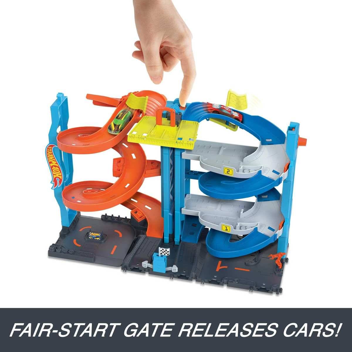 Hand zeigt auf ein Spielzeug-Hot-Wheels-Rennstreckenset mit orange- und blauem spiralförmigen Rampen; die Bildunterschrift sagt „FAIR-START-TOR GIBT AUTOS FREI!