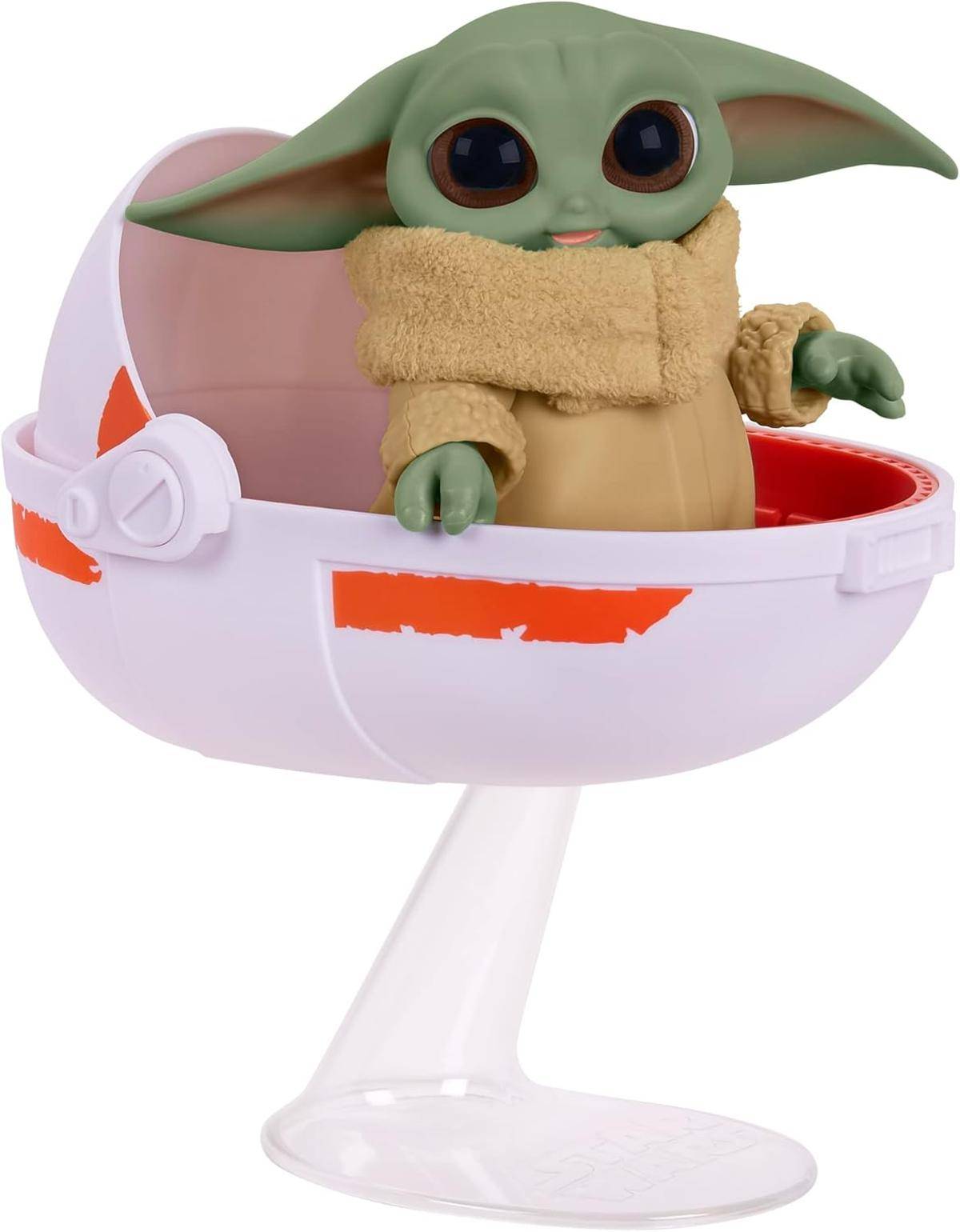 Grogu-Babyfigur, sitzend in einem weißen podförmigen Schwebewagen auf einem durchsichtigen Ständer, bekleidet mit einem beigen Mantel und Schal.