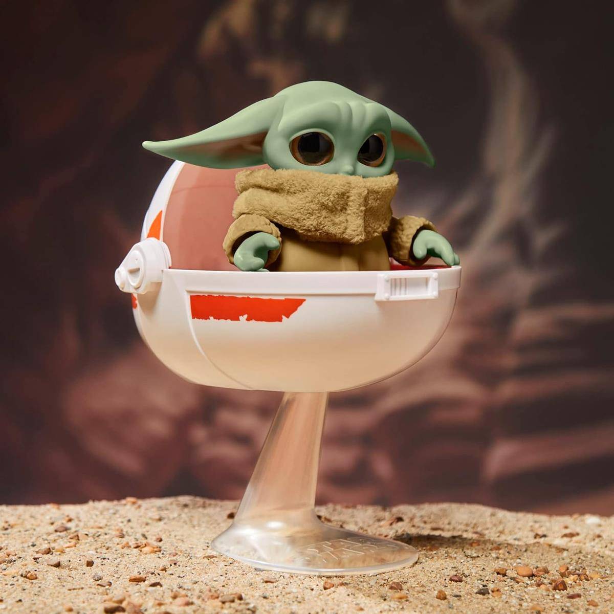 Kleine grüne Figur, die Baby Yoda ähnelt, sitzt in einer weißen, eiförmigen Kapsel auf einem Ständer vor einem verschwommenen Hintergrund.