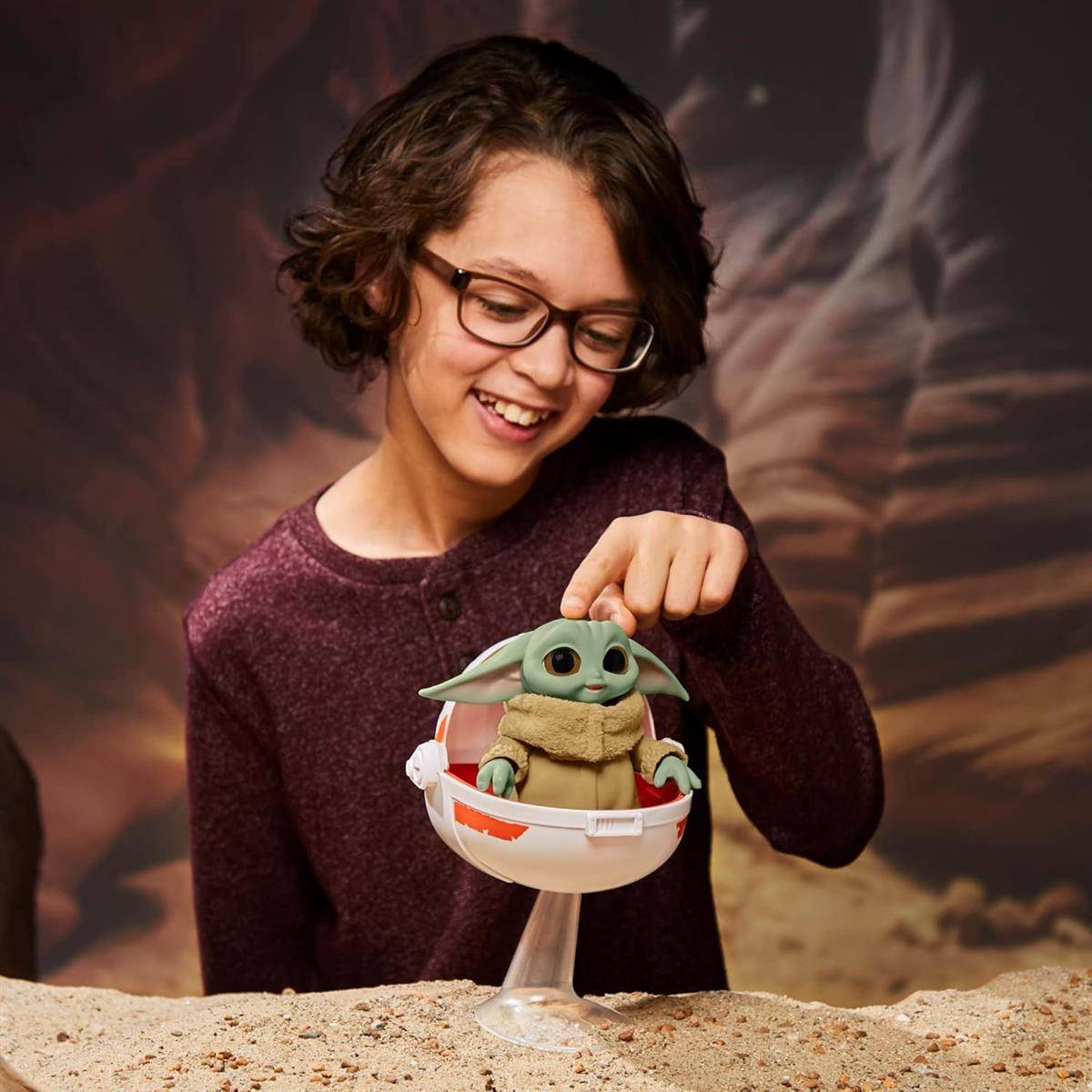 Kind mit Brille, das lächelnd ein Baby-Yoda-Spielzeug in einer schwebenden Kapsel gegen einen Star-Wars-thematisierten Hintergrund hält.