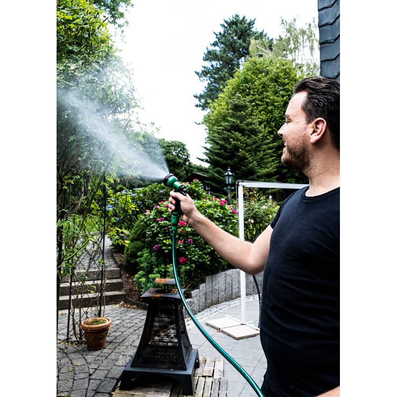Eine Person gießt Pflanzen in einem Garten mit einem Gartenschlauch. Grünpflanzen und Gartenmöbel sind zu sehen, und die Person trägt ein schwarzes Shirt.