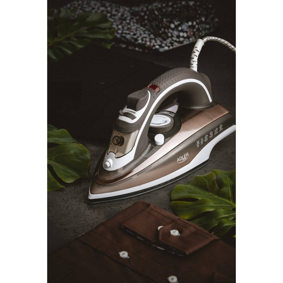 Ein Bügeleisen auf einer dunklen Oberfläche neben einem braunen Hemd und grünen Blättern, was auf eine Umgebung zum Bügeln von Kleidung hindeutet.