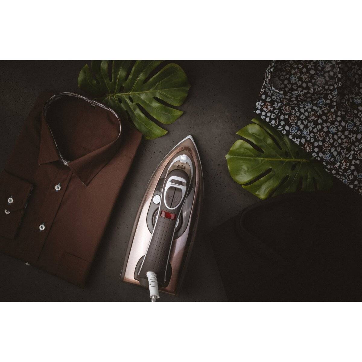 Ein ordentlich gefaltetes braunes Hemd liegt neben einem modernen Dampfbügeleisen auf einer dunklen Oberfläche mit zwei tropischen Blättern und blumig gemustertem Stoff.