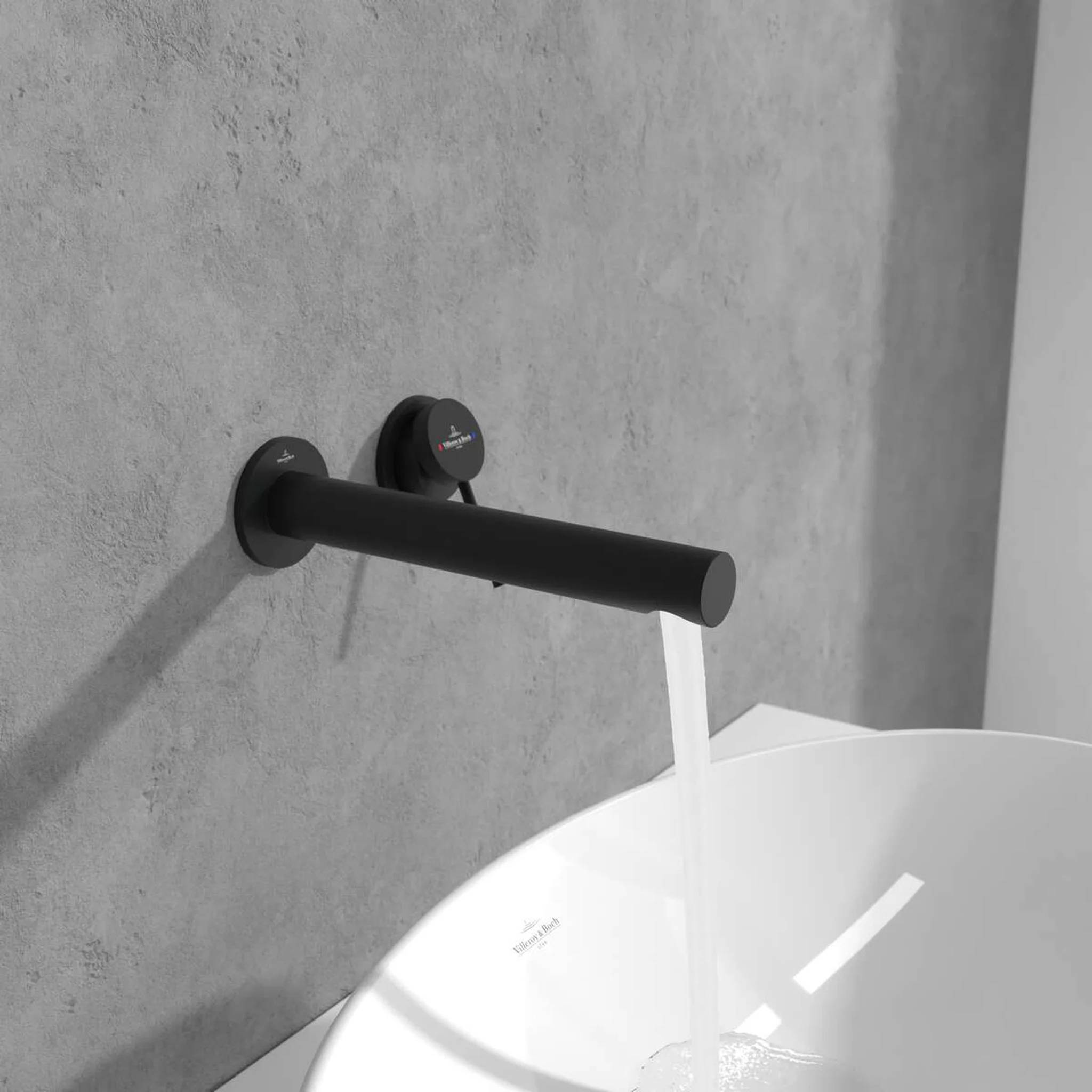 Schwarzer wandmontierter Wasserhahn, der Wasser in ein weißes rundes Waschbecken vor einer graufliesigen Wand gießt.