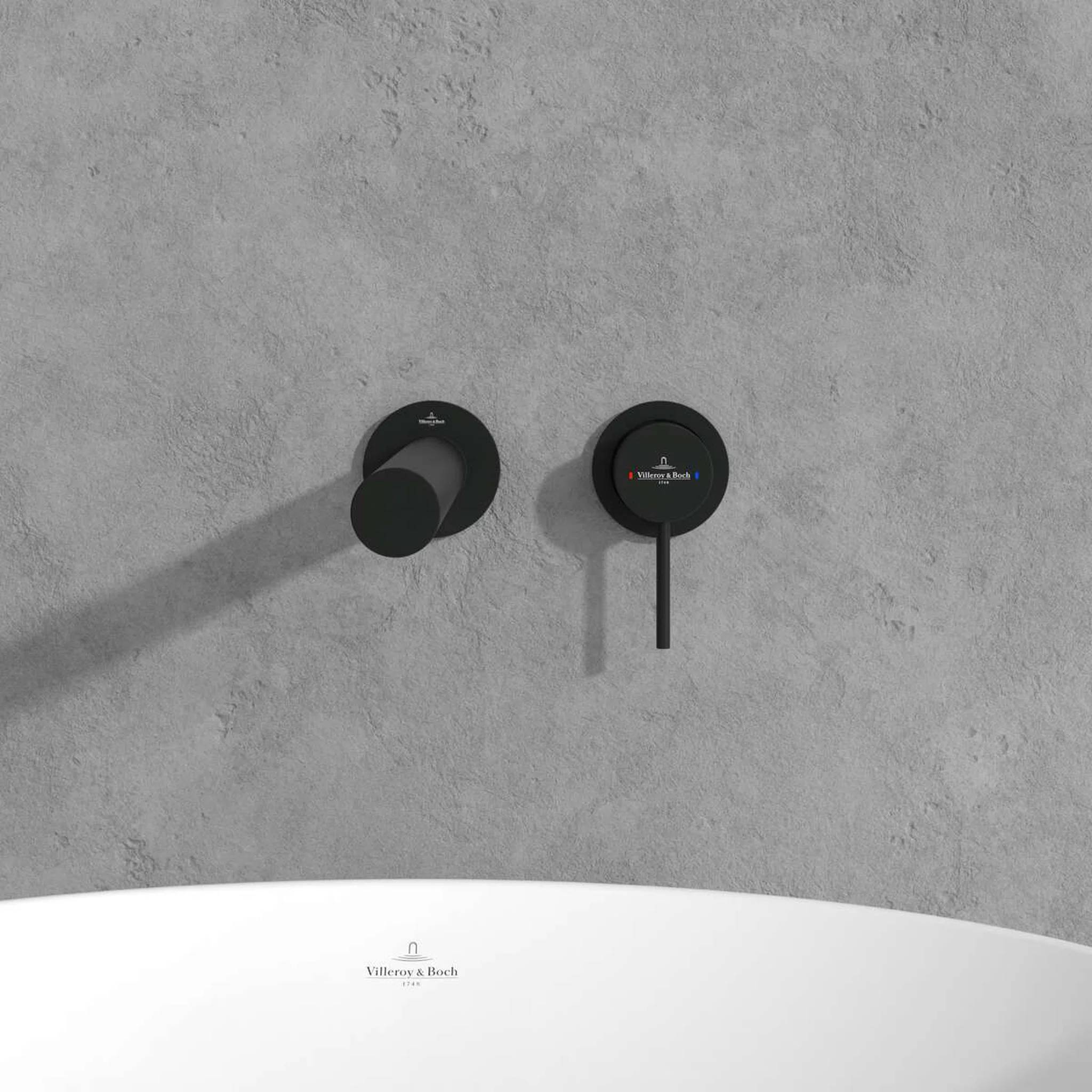 Zwei mattschwarze, wandmontierte Badezimmerarmaturen über einem weißen Waschbecken vor einer graustrukturierten Wand.