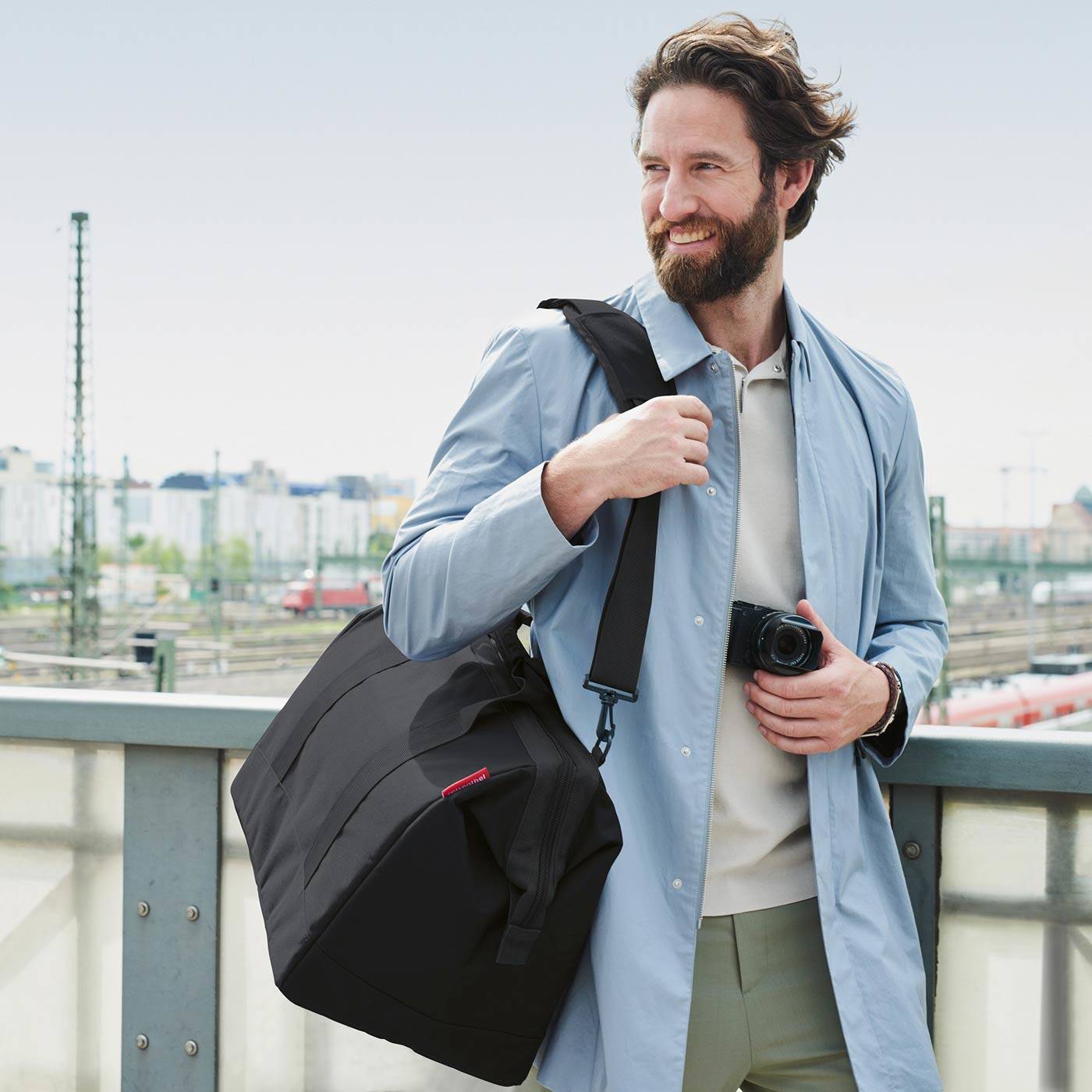 Lächelnder Mann im Freien auf einer Brücke, trägt einen hellblauen Mantel und trägt eine große schwarze Tasche mit einer Kamera in der Hand.