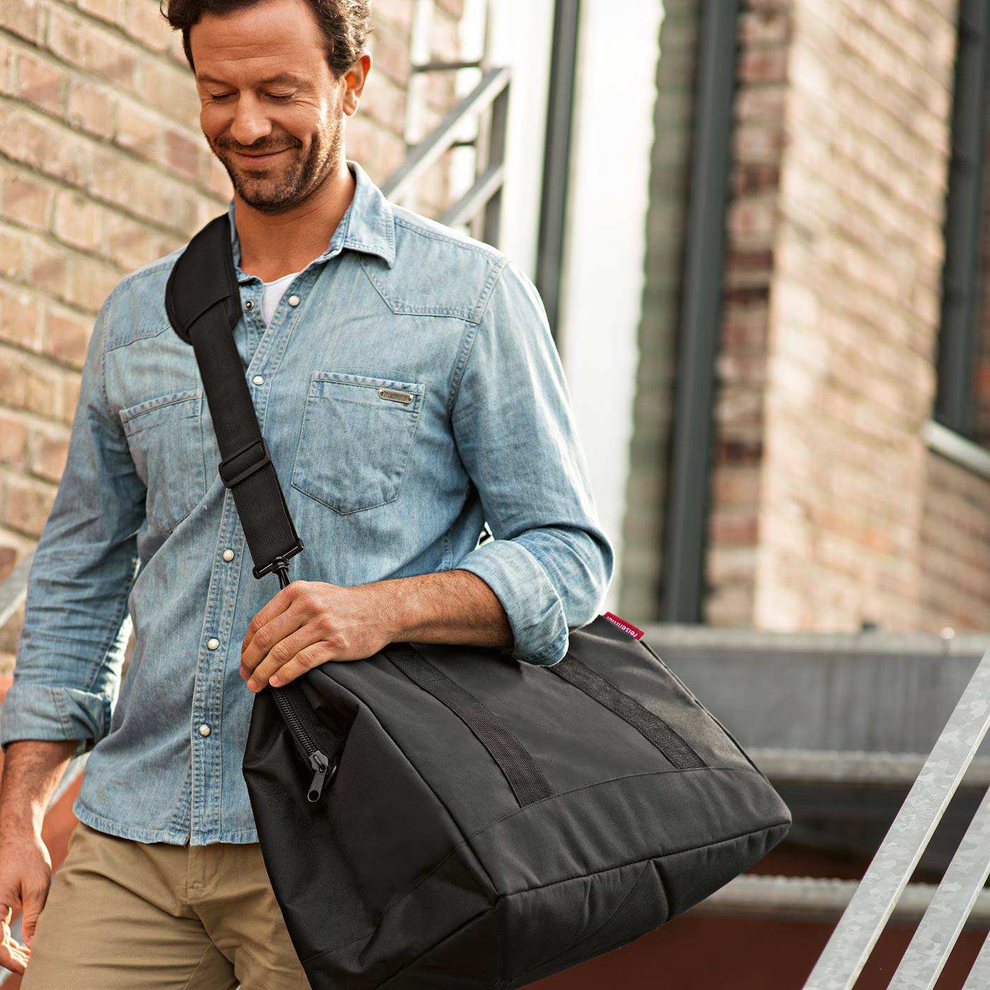 Mann mit einer schwarzen Schulter-Reisetasche im Freien neben einem Ziegelgebäude, trägt ein Jeanshemd und Khaki-Hose.