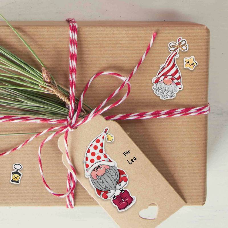 Ein eingepacktes Geschenk mit rot-weißer Schnur, einem Tannenreis und einem Anhänger mit einer Gnom-Illustration. Festliche Sticker mit Gnomen und Sternen schmücken das Paket.