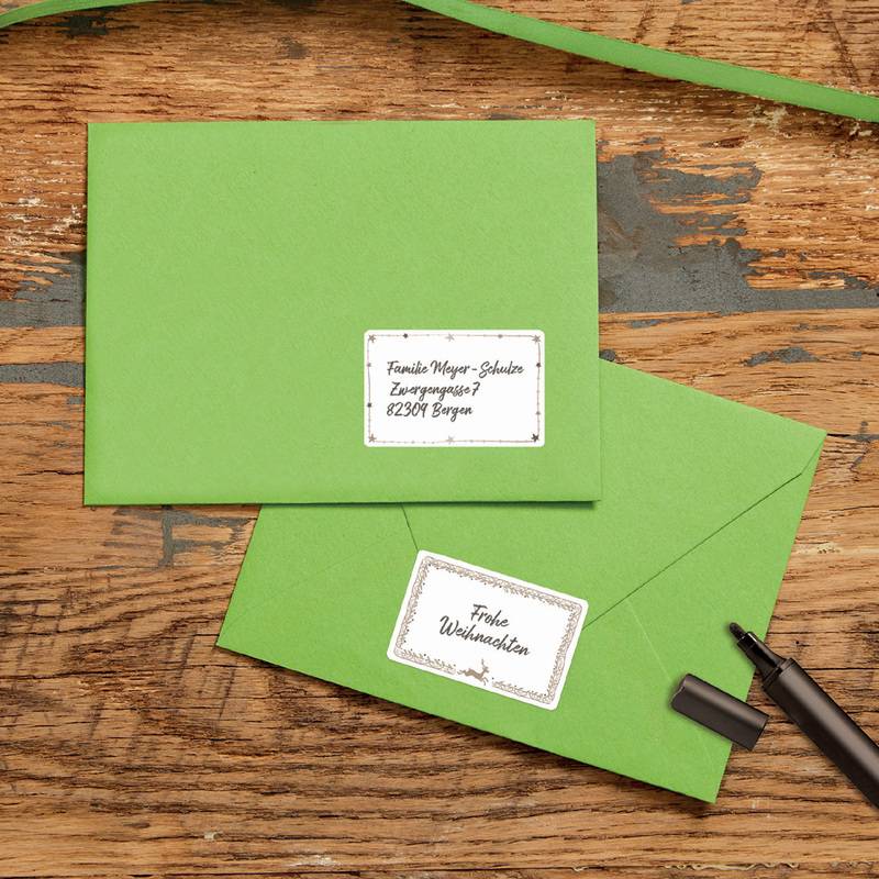 Zwei grüne Briefumschläge liegen auf einem Holztisch, einer davon mit einem sichtbaren Adressetikett, auf dem steht: 'Frieda Meier-Schulze, Hauptstraße 5, 12345 Berlin'.