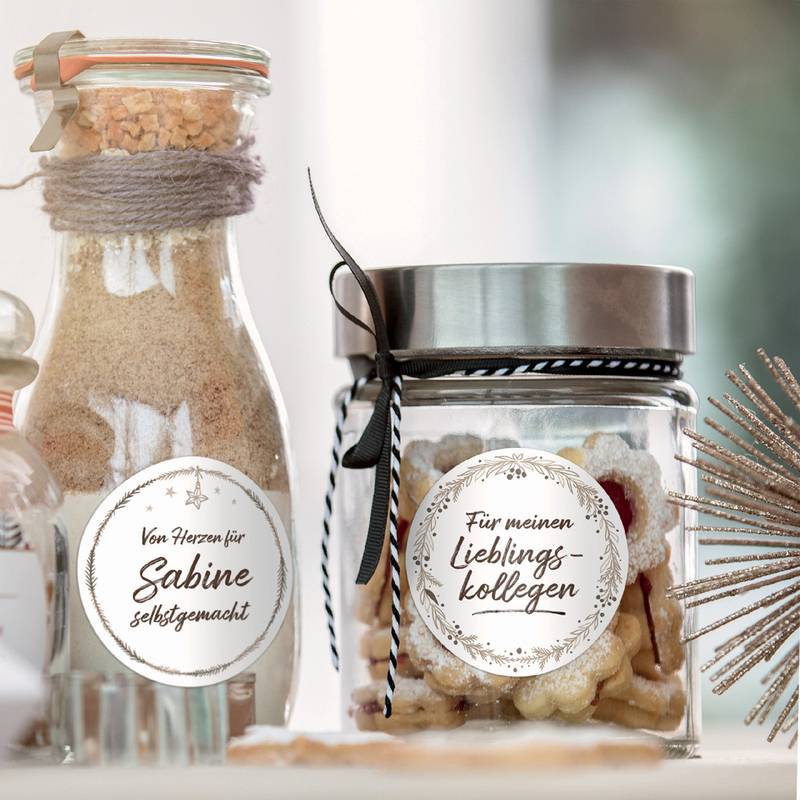 Zwei Gläser mit Etiketten. Das linke Glas trägt die Aufschrift ‚Sabine selbstgemacht', gefüllt mit einer sandartigen Substanz. Das rechte Glas, ‚Für meinen Lieblingskollegen', enthält sternförmige Kekse. Beide sind festlich dekoriert.