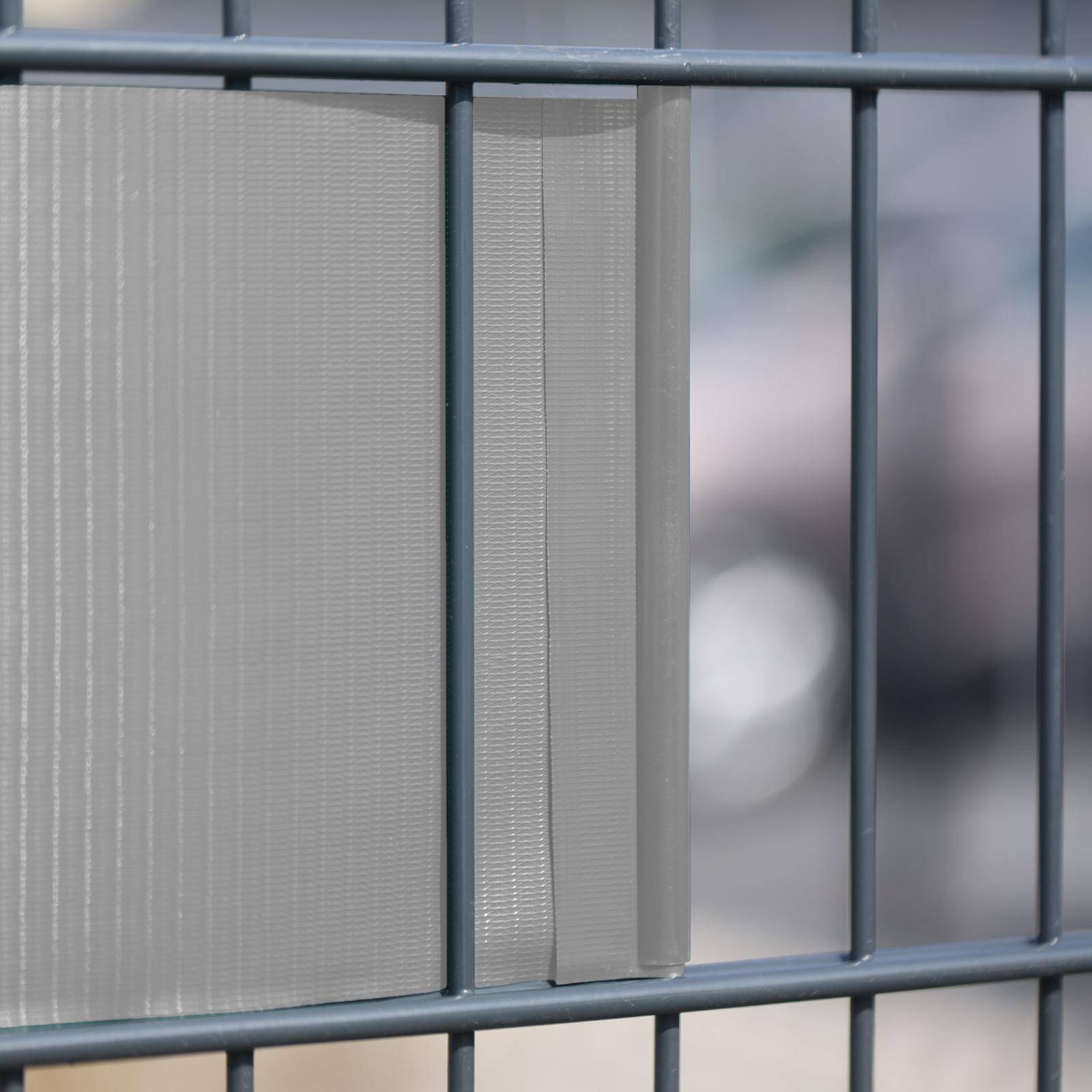 Nahaufnahme eines grauen Metallzauns mit vertikalen Stäben und durchscheinenden Sichtschutzpanelen, mit unscharfem Hintergrund dahinter.