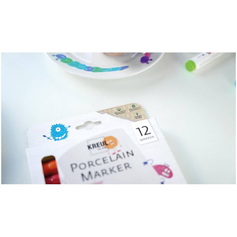 Eine 'Porzellan-Marker'-Packung mit 12 Markern liegt teilweise geöffnet auf einem weißen Tisch. In der Nähe befindet sich ein Pappteller mit bunten Farbstrichen.