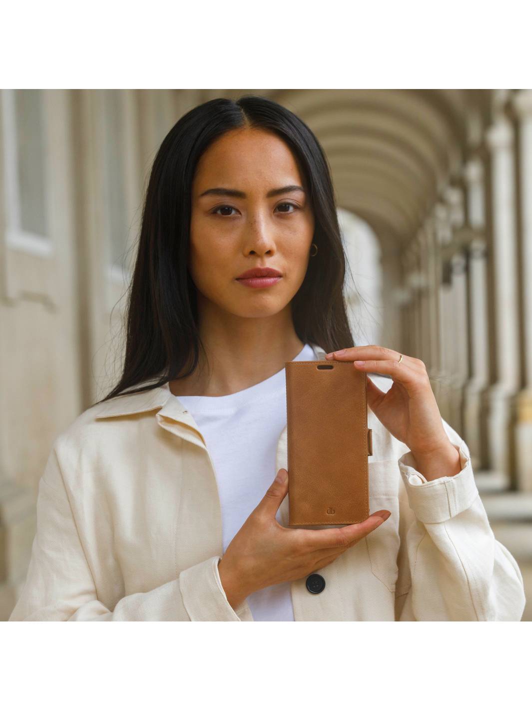 Porträt einer Frau in einem cremefarbenen Mantel, die eine braune Smartphone-Hülle oder ein Portemonnaie hochhält, stehend in einem gewölbten Außenflur.