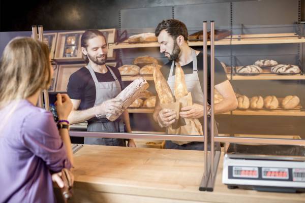 Zwei Bäcker an einem Tresen unterhalten sich über Laibe Brot in einer Bäckerei, die mit verschiedenen Brotsorten gefüllt ist, die auf Regalen hinter ihnen ausgestellt sind.