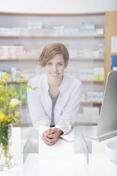 Ein Apotheker in einem weißen Kittel lächelt hinter einem Schalter mit einer Schutzvorrichtung und ist umgeben von Regalen mit Medikamenten und einem Computer.