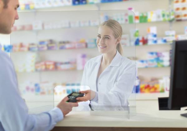 Apotheker in einem weißen Kittel lächelt hinter einem Apothekentresen, während ein Kunde mit einer Kreditkarte bezahlt.