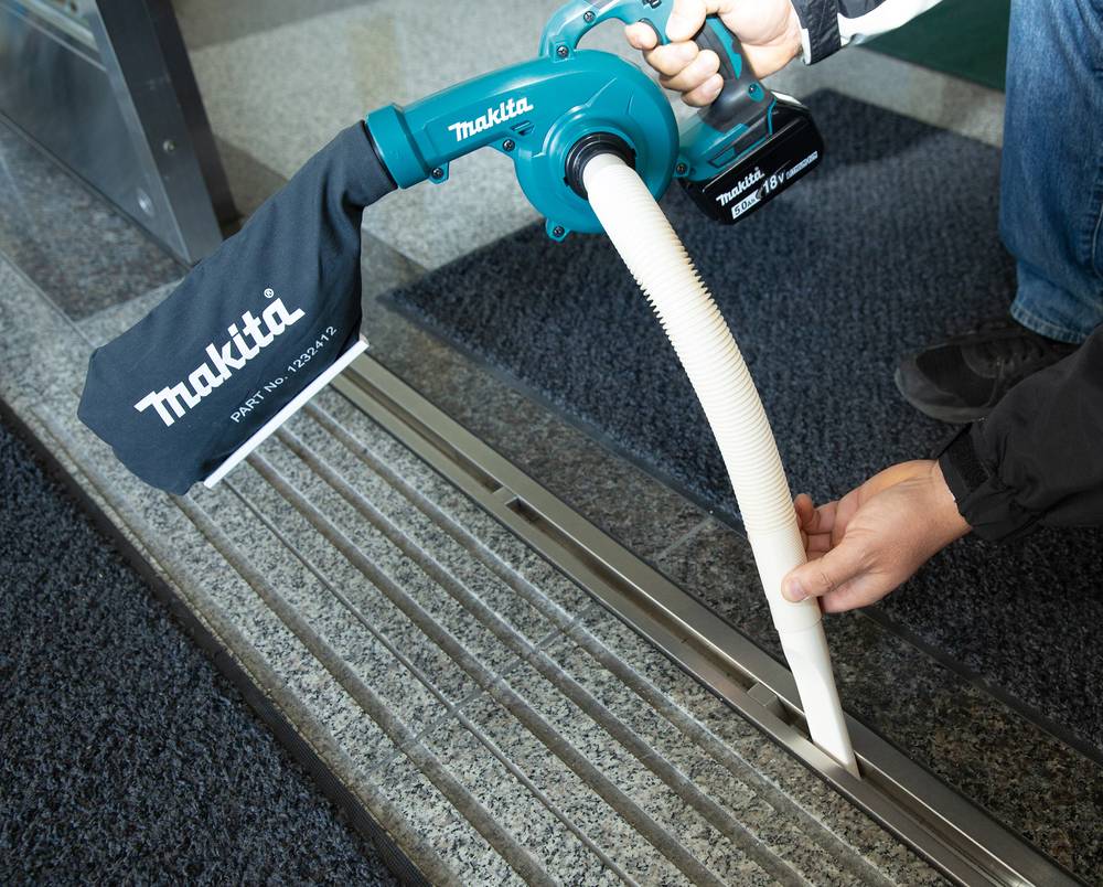 Person, die eine Makita-Staubsauger-Aufsatz verwendet, um Verschmutzungen von einem Bodenablauf-Gitter in Innenräumen zu entfernen.