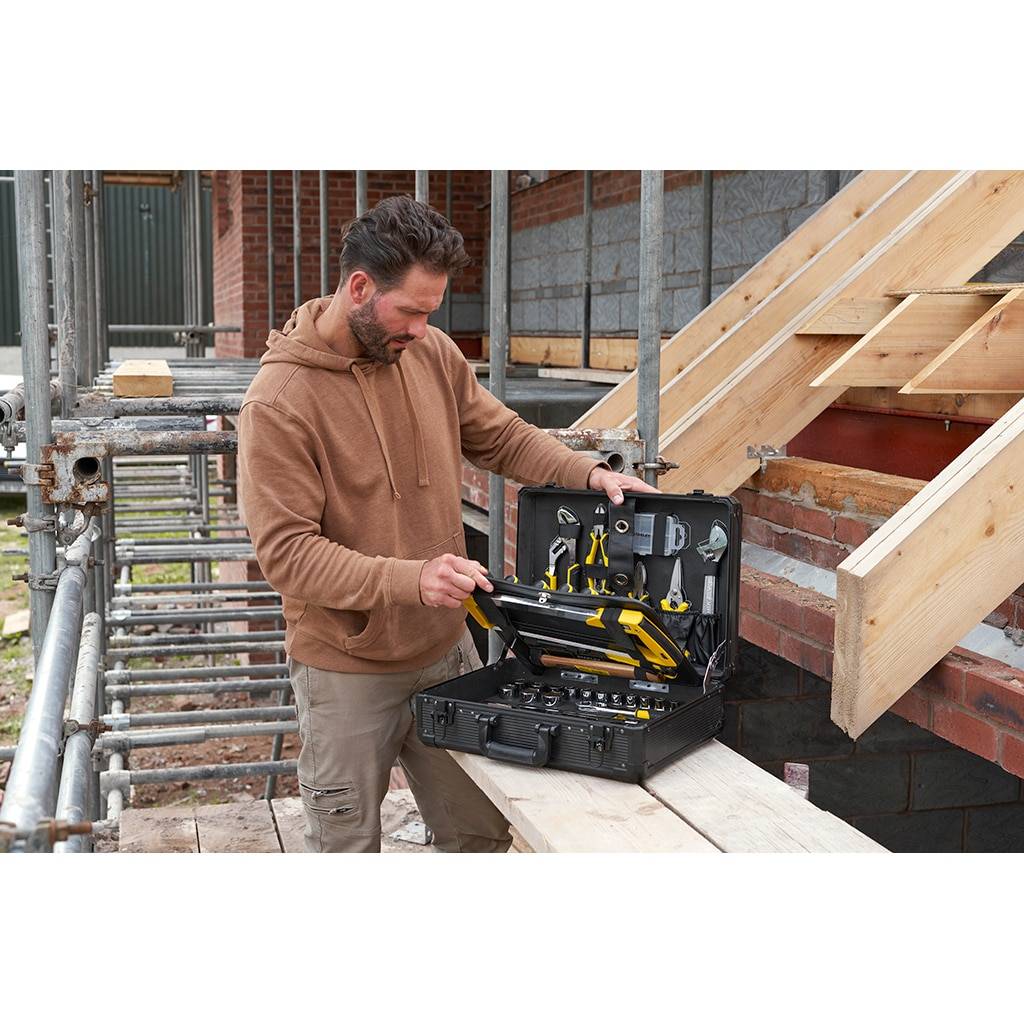 Bauarbeiter inspiziert einen Werkzeugkasten mit Handwerkzeugen neben Ziegelmauerwerk und Holzdachkonstruktion auf einer Baustelle.