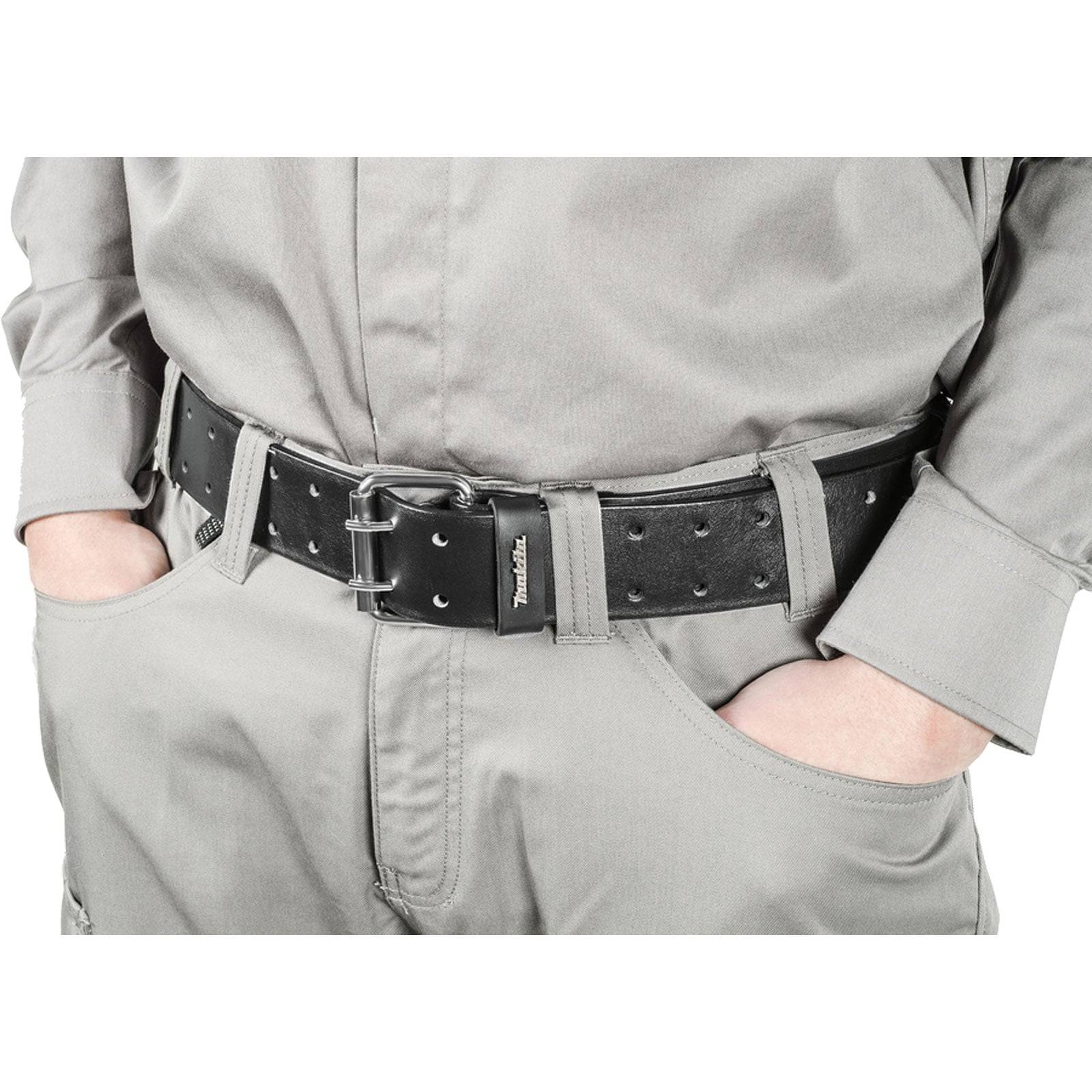 Eine Person, die eine graue Uniform trägt, mit den Händen in den Taschen, wobei ein schwarzer Ledergürtel mit Metalldetails zu sehen ist.