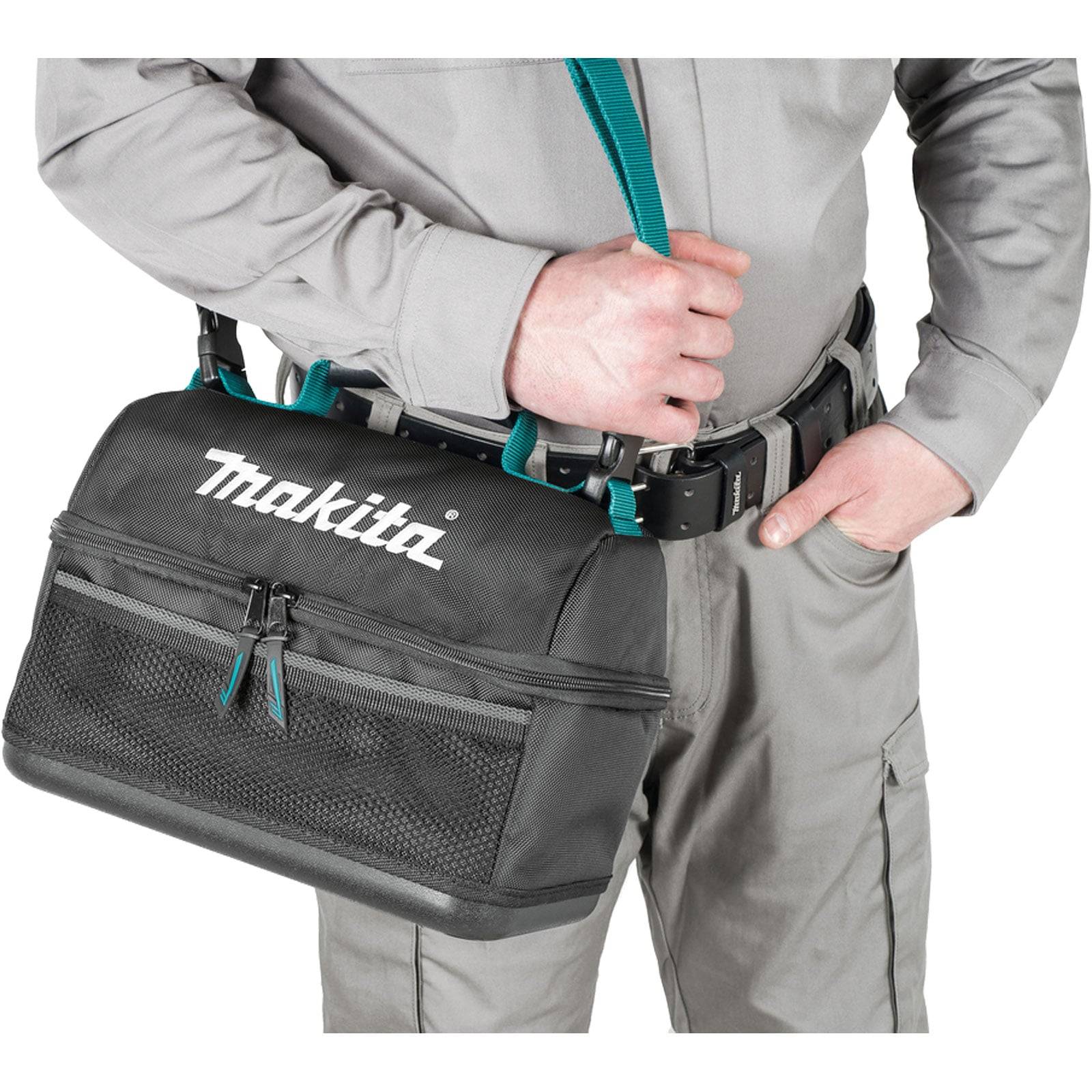 Eine Person in einem grauen Outfit hält eine schwarze Makita-Werkzeugtasche mit Schultergurt und Reißverschlusskompartimenten, was Arbeitsbereitschaft andeutet.