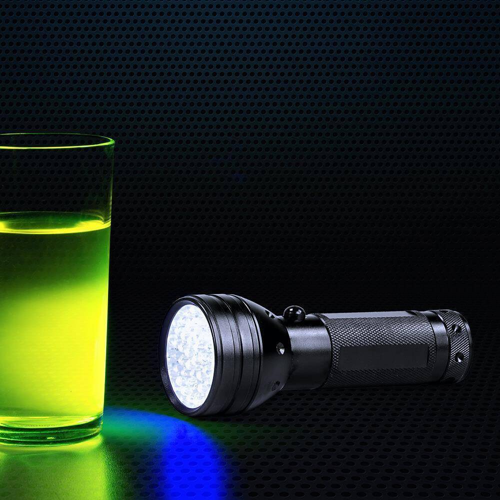 Schwarze Taschenlampe auf einer dunklen Oberfläche neben einem Glas mit leuchtend grüner Flüssigkeit, wobei der Strahl blaue und grüne Lichtreflexionen wirft.