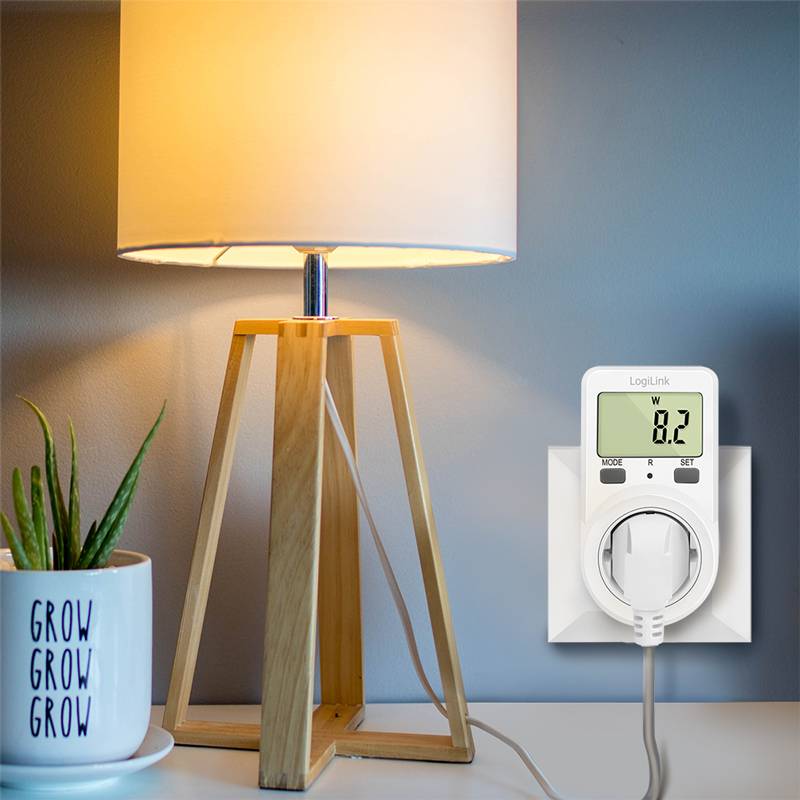 Eine Tischlampe aus Holz leuchtet neben einem Stromzähler, der in eine Steckdose gesteckt ist und '0,2W' anzeigt. Daneben steht eine Pflanze in einem Topf mit der Aufschrift 