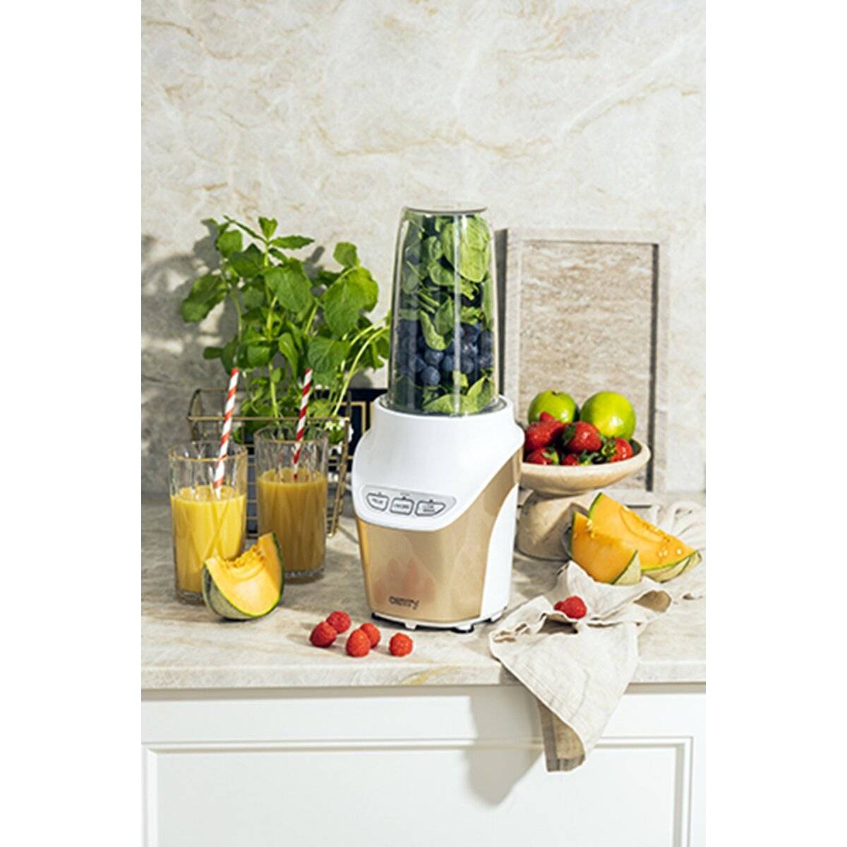 Ein Mixer gefüllt mit Spinat und Blaubeeren auf einer Küchentheke, umgeben von Gläsern Orangensaft, Melonen, Äpfeln und Erdbeeren.