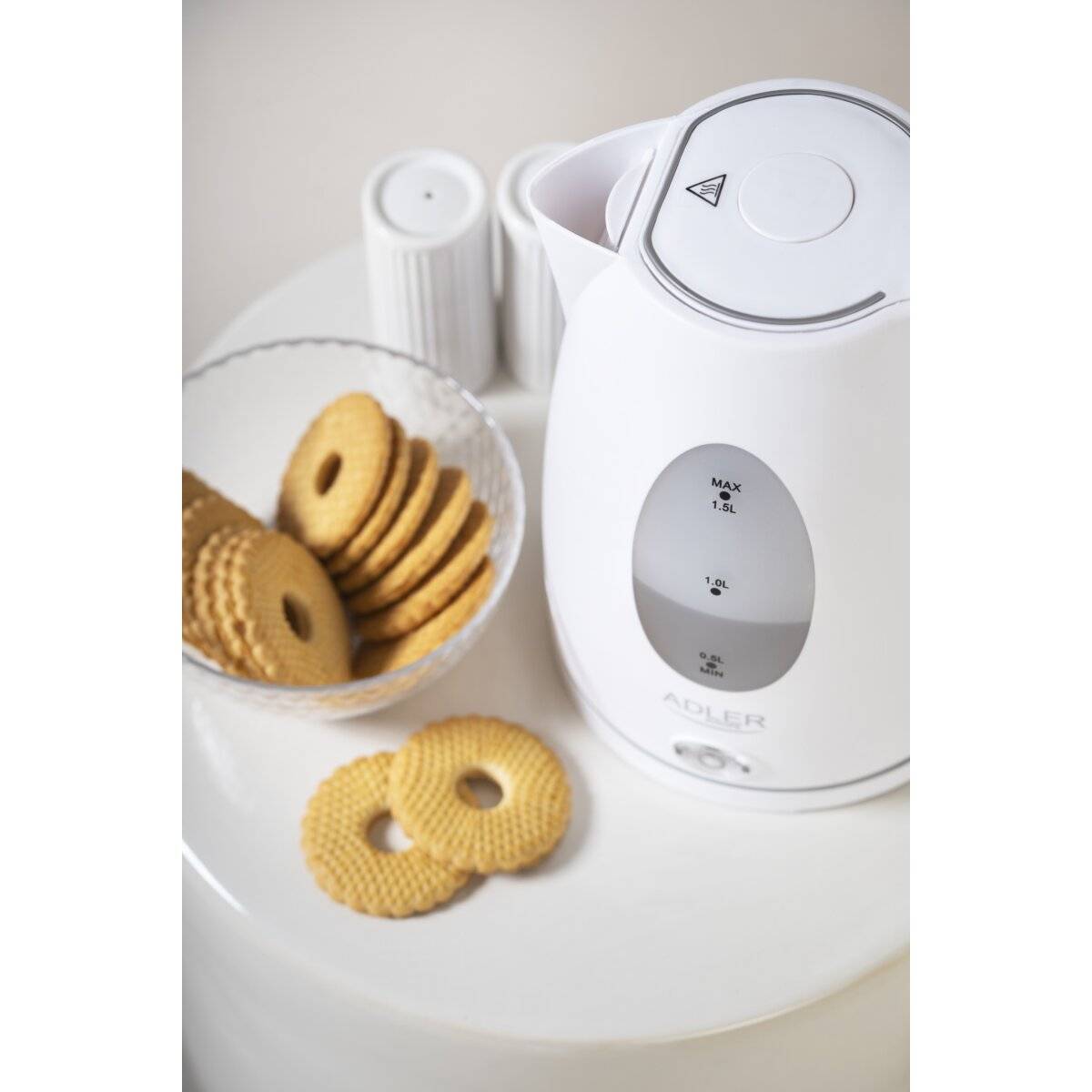 Ein weißer Wasserkocher neben einer Schale mit runden Butterkeksen auf einer weißen Oberfläche. Der Wasserkocher hat eine sichtbare Wasserstandsanzeige.