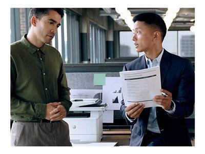 Zwei Personen in einem Büro, die in der Nähe eines Druckers stehen. Eine Person hält ein Dokument in der Hand. Sie scheinen sich zu unterhalten.